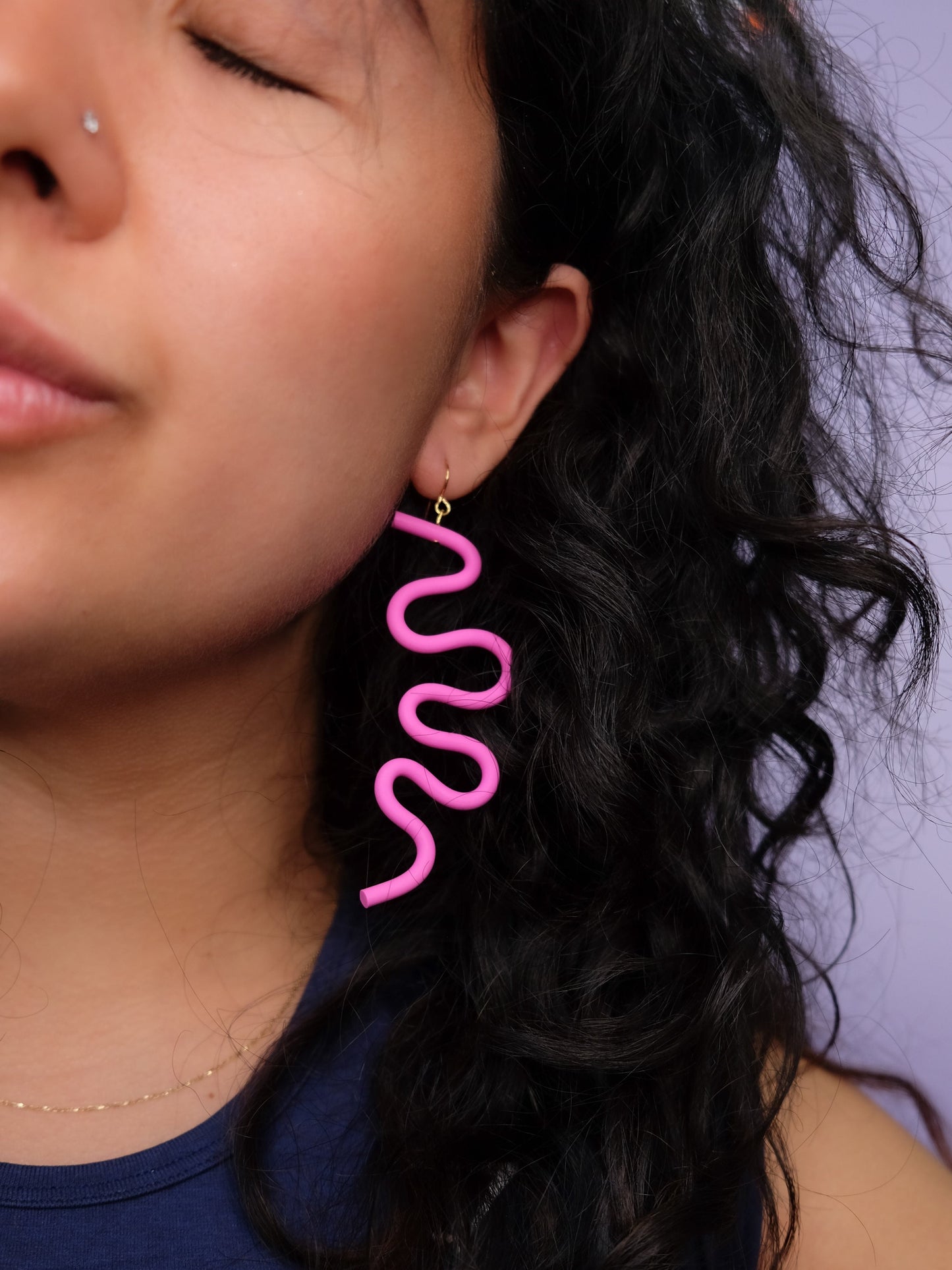 Close-up of a person wearing pink wavy earrings with a blurred, bold, lightweight, sculptural, squiggle-shaped statement piece #color_pink