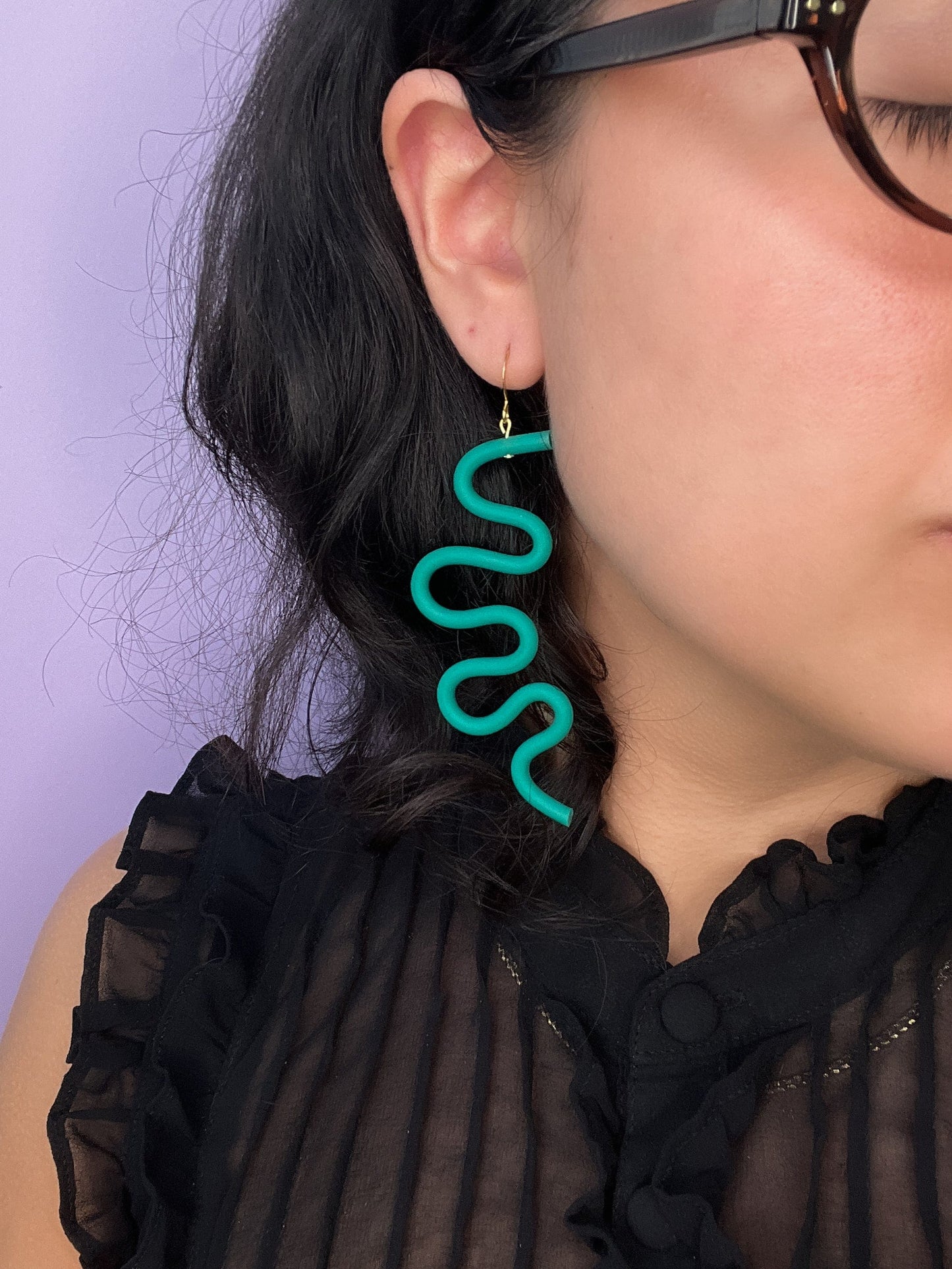Emerald wavy earring, oversized, matte, tubular, sculptural, worn by a person with dark hair and glasses #color_emerald 